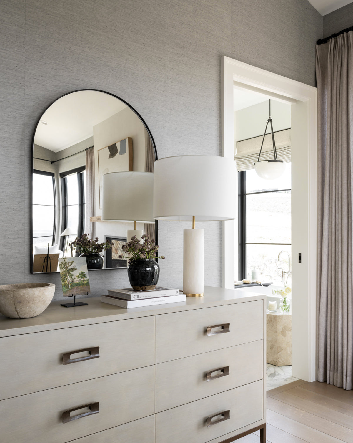 A modern dresser holds a white lamp, books, a bowl, and a vase with flowers. The Wylie Arched Mirror with shatterproof glass reflects windows and wall art as sunlight streams in through an open doorway and curtained window, by Vendixs