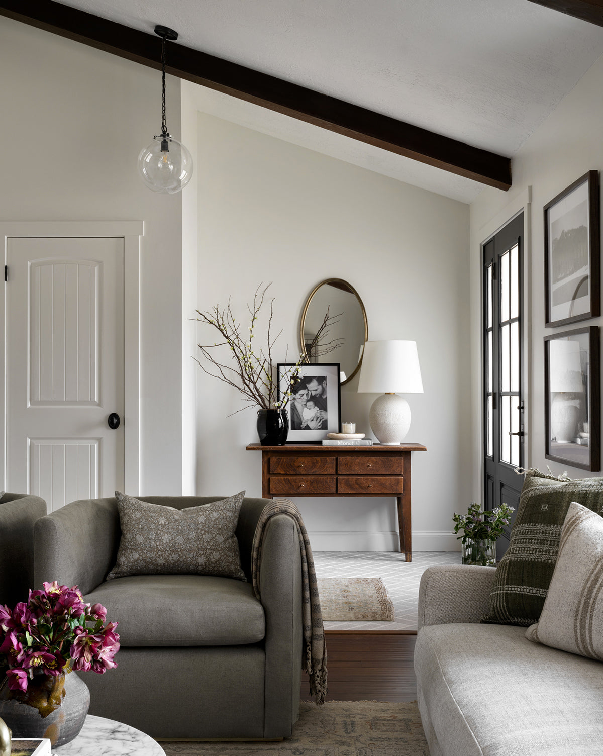 A cozy living room with neutral tones features a gray armchair, sofa, round marble coffee table with purple flowers, wooden console under a round mirror, and is illuminated by the Visual Comfort & Co. Signature Katie Globe Pendant—a setting reminiscent of the timeless elegance found at Vendixs