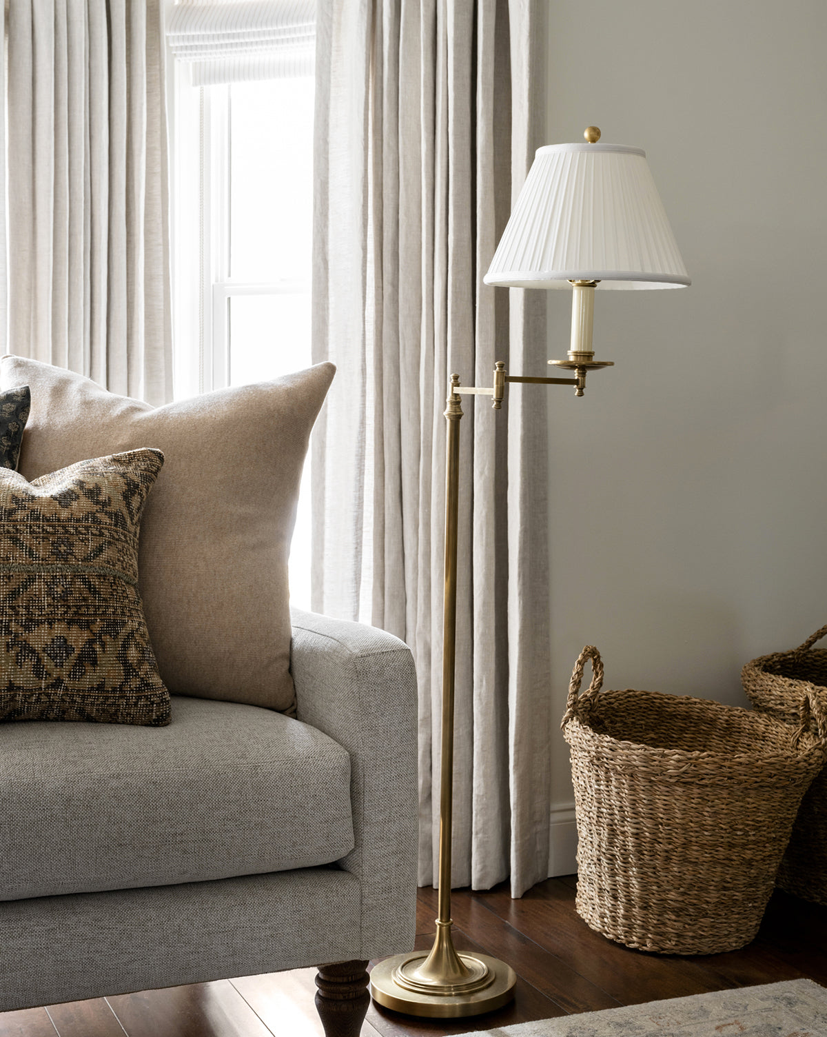 The Dorchester Swing Arm Floor Lamp with a pleated silk shade stands beside a light gray sofa topped with patterned and solid throw pillows. Two woven baskets sit on the wooden floor near beige curtains by the window, by Vendixs