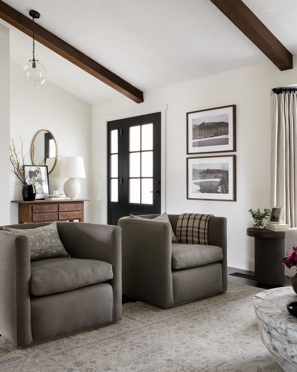 A cozy living room features two Reese accent chairs side-by-side with down-wrapped cushions, a handcrafted alder wood console table topped with a lamp and mirror, framed art on white walls, exposed ceiling beams, and a black front door with glass panels by McGee & Co.