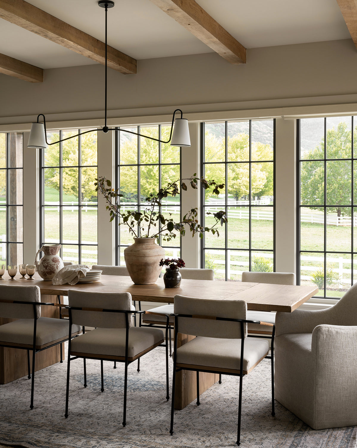 A modern dining room featuring the Dillon Extension Dining Table with six chairs, a light gray armchair, a large ceramic vase with branches, and large windows overlooking trees and a white fence by McGee & Co.