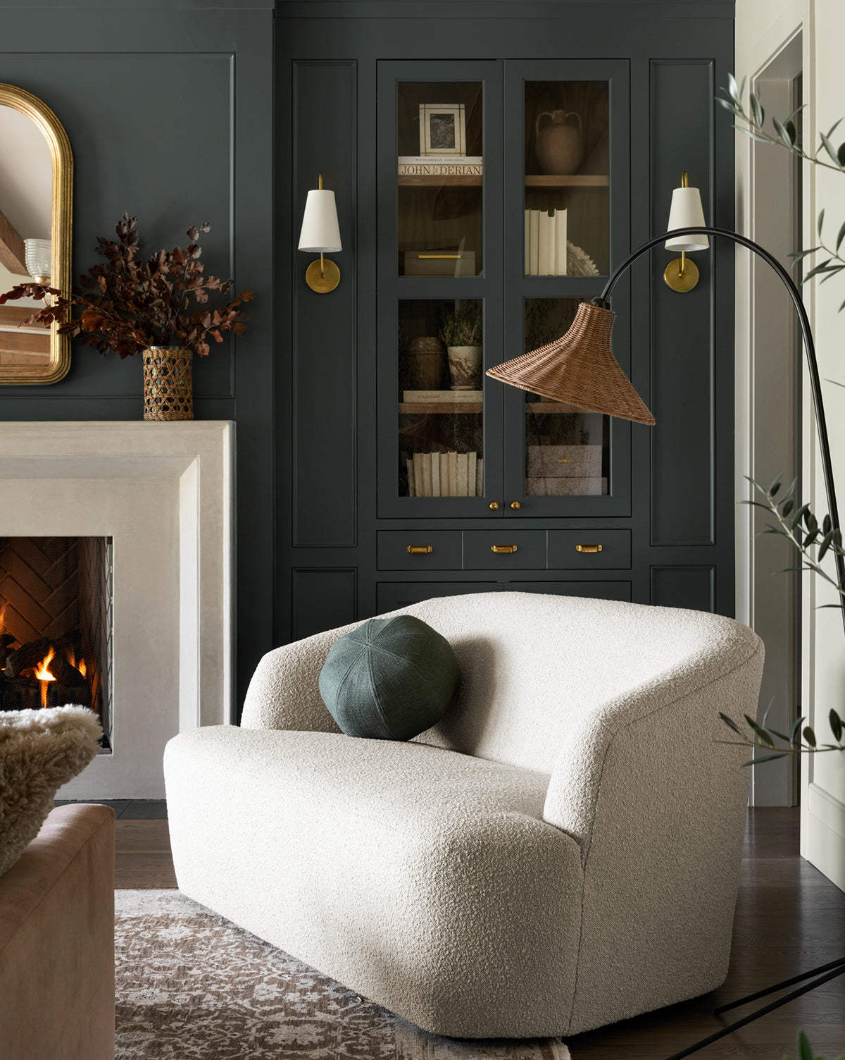 A cozy living room with a cream boucle loveseat, round green pillow, the Johanson Woven Floor Lamp, built-in dark cabinets with brass hardware, a lit fireplace, and a decorative mirror above the mantel by Vendixs