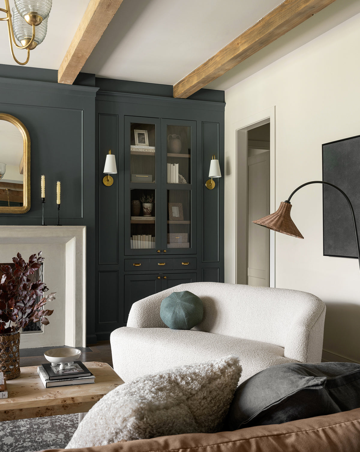 A modern living room features the Makerspalm Byron Settee 68 with a curved back, a wooden coffee table, and built-in dark cabinets with glass doors. Decorative objects, books, and soft lighting add to the cozy, stylish space. Vendixs