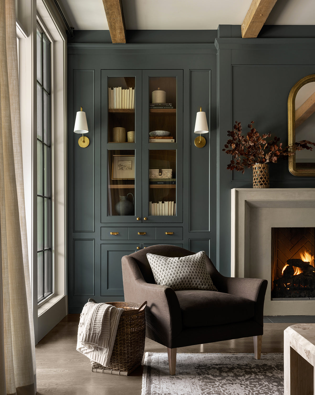 A cozy living room features a Prudence Lounge Chair with a classic silhouette and patterned pillow, a woven basket with a blanket, built-in bookcase, crackling fireplace, and elegant wall sconces—all in a warm, inviting setting by McGee & Co.
