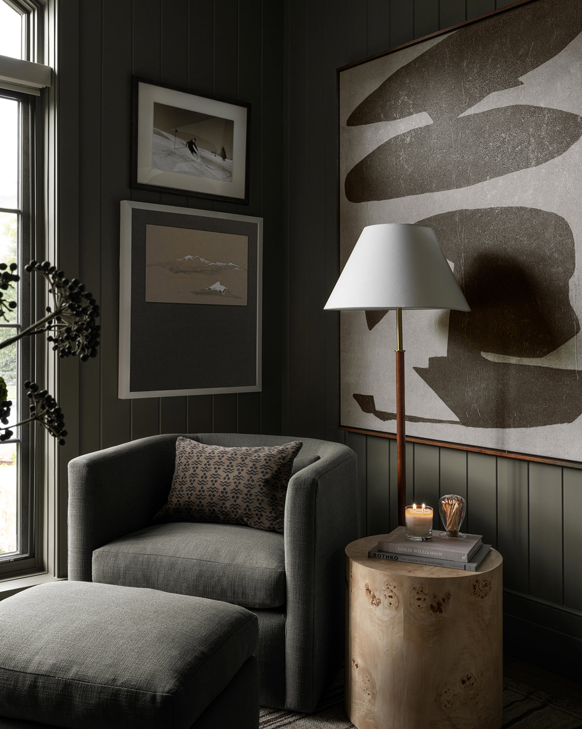 A cozy modern living room corner with a Reese accent chair in dark grey featuring down-wrapped cushions, a matching ottoman, a small round handcrafted alder wood side table holding a candle and glass, abstract art, and dark-paneled walls near a window, by McGee & Co.