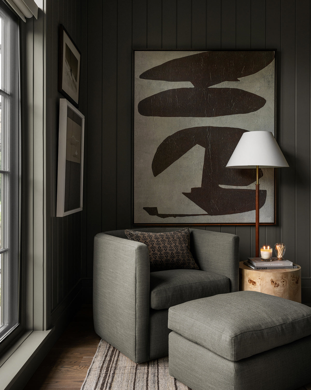 A cozy reading nook with a gray armchair, the Reese Ottoman, a patterned pillow, and a side table. A modern lamp and abstract artwork decorate the dark-paneled wall beside a large window filled with soft natural light, by McGee & Co.
