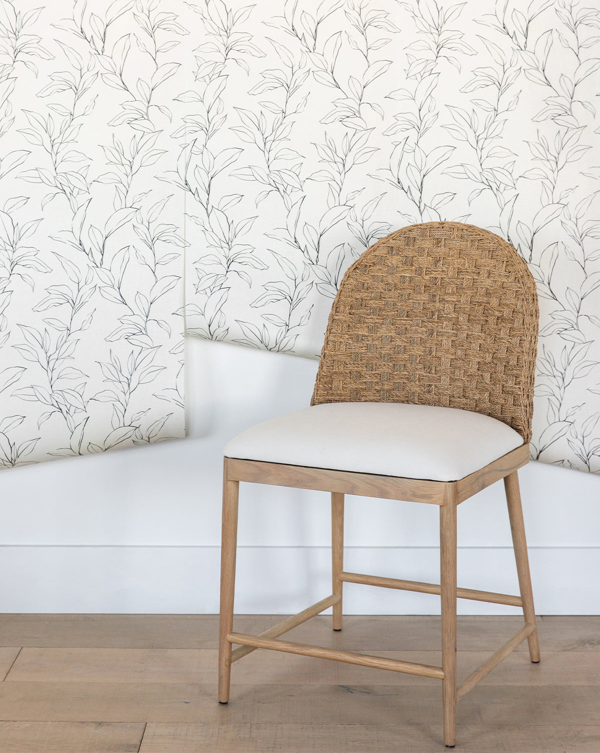 A wooden chair with a woven rattan backrest and cream cushion sits before a white wall featuring Cecile Floral Print Wallpaper by York Wallcoverings, its light leafy pattern partially peeling at the bottom—a vignette that feels straight from Vendixs