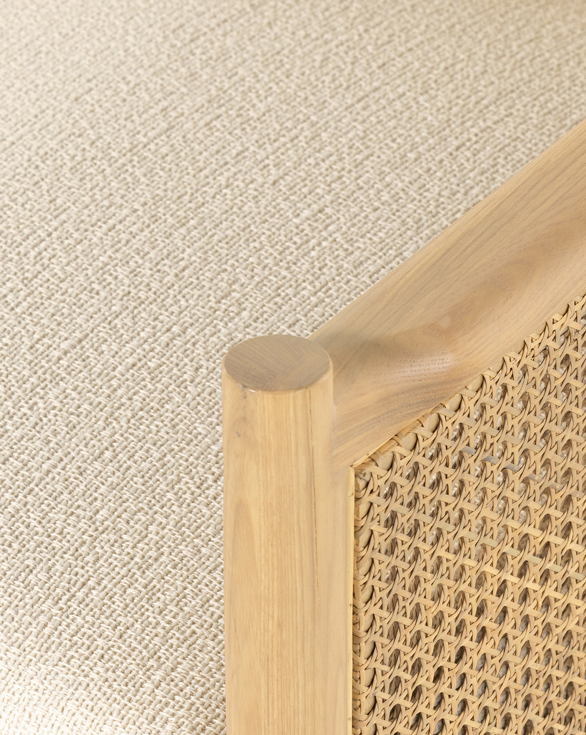 Close-up of the Channing Lounge Chair, featuring a light wood frame and woven rattan panel inspired by Parisian design. The corner post and woven detail stand out, with the chair set on a textured beige fabric surface, by Vendixs
