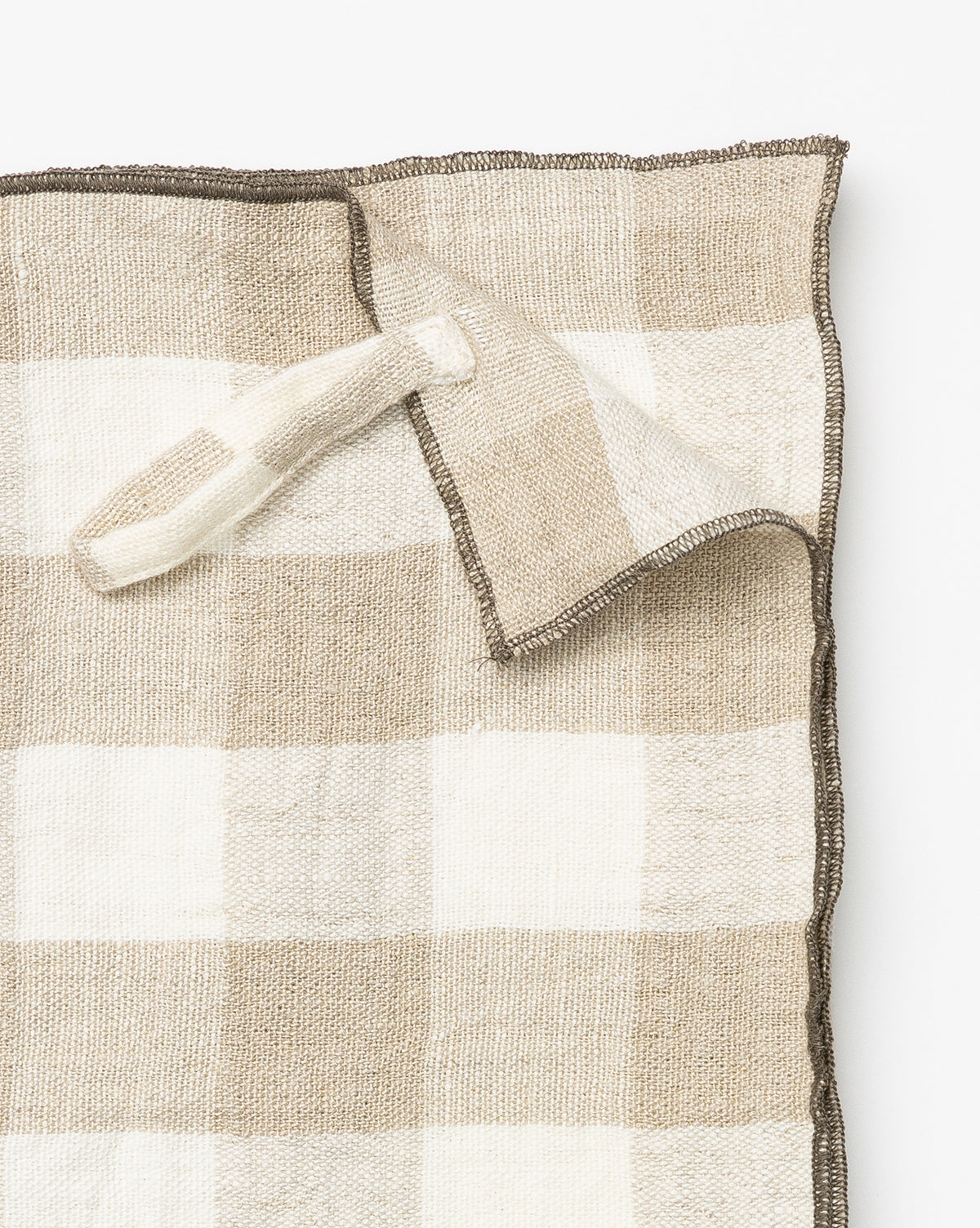 The Charlie Check Tea Towel features a beige and white checkered linen design with a hanging loop, shown partially folded at one corner—ideal for elegant table settings against a white background, by Vendixs