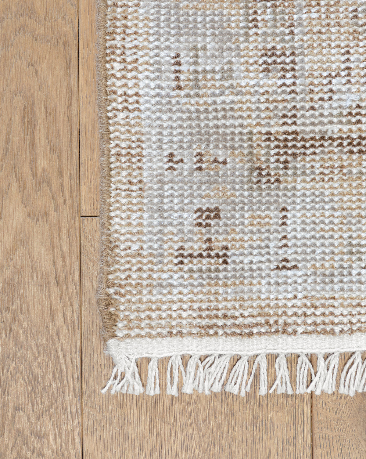 A close-up of the Charlot Hand-Knotted Rug’s corner shows its white fringe and textured beige, brown, and white pattern on a light wooden floor, by Vendixs