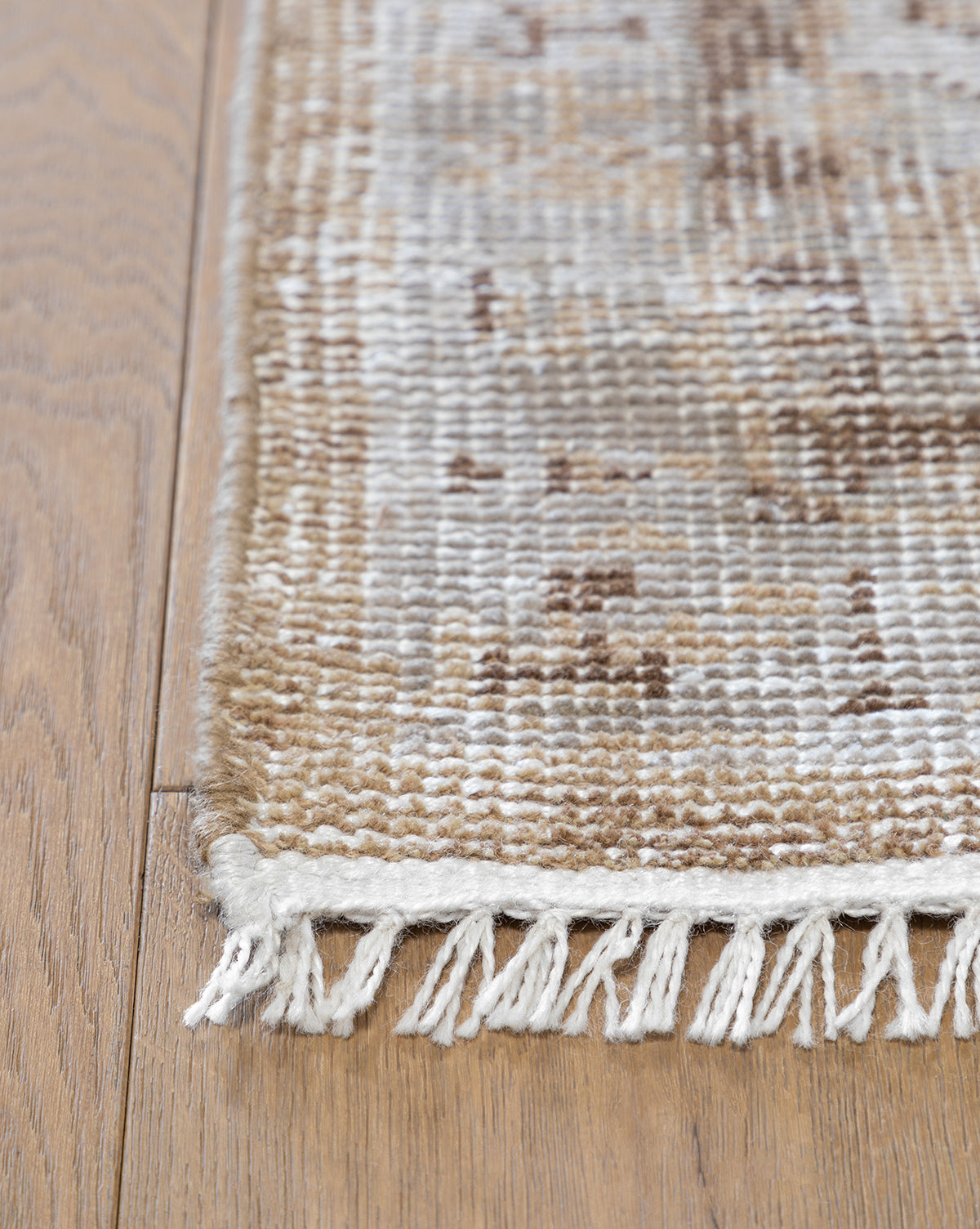 A close-up shows the corner of a Charlot Hand-Knotted Rug in beige, brown, and cream with white fringe, placed on a light wood floor, by Vendixs