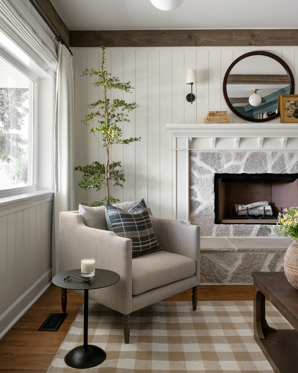 A cozy living room corner features the Gemma Lounge Chair in high-quality fabric, a plaid pillow, a small round black side table with a candle, stone fireplace, round mirror above the mantel, and a tall plant by white-curtained windows by McGee & Co.
