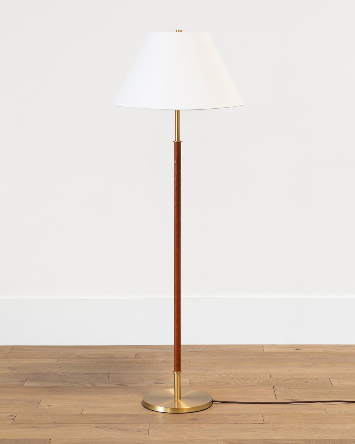 The Elijah Floor Lamp has a white conical shade, slim brass pole, and gold base, shown on a light wood floor with a plain white wall background from Vendixs