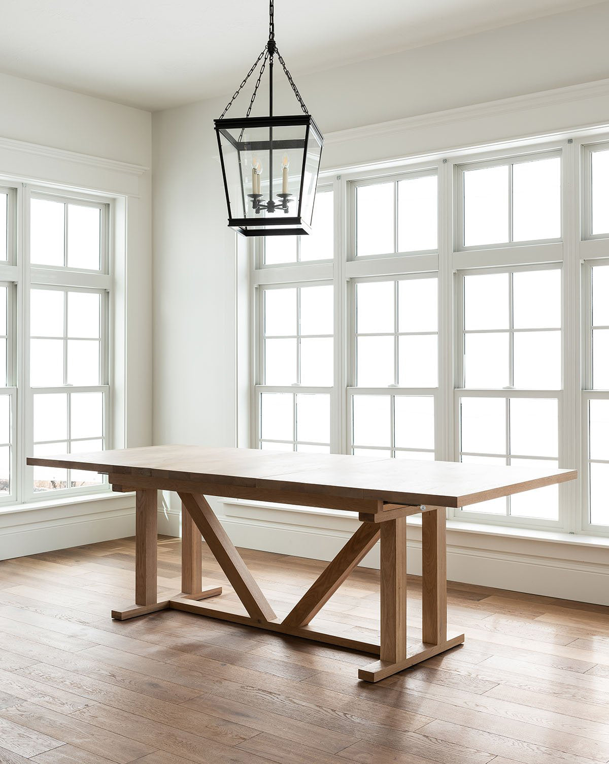 A minimalist room with large, white-framed windows features the Emory Extension Dining Table with a trestle base, paired with a black metal lantern pendant light hanging above on a wooden floor, by McGee & Co.