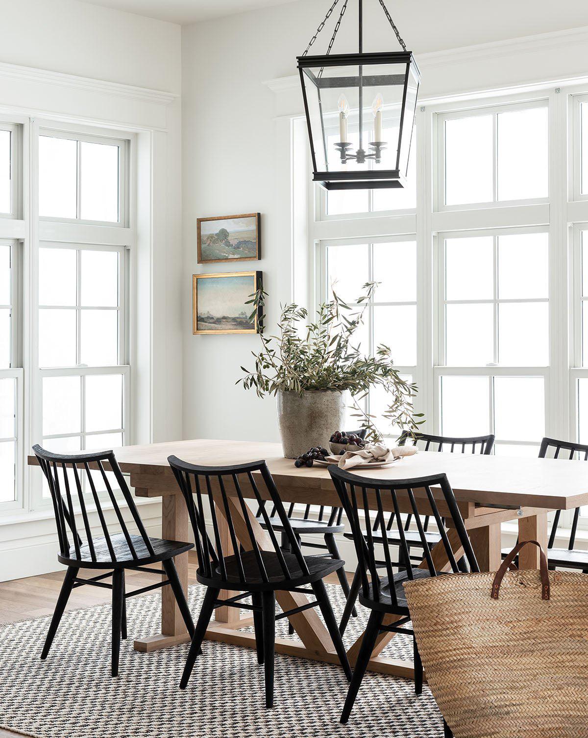 Bright dining area with large windows, an Emory Extension Dining Table, black chairs, potted plant centerpiece, woven rug, two landscape paintings on the wall, and a lantern-style pendant above the table, by Vendixs
