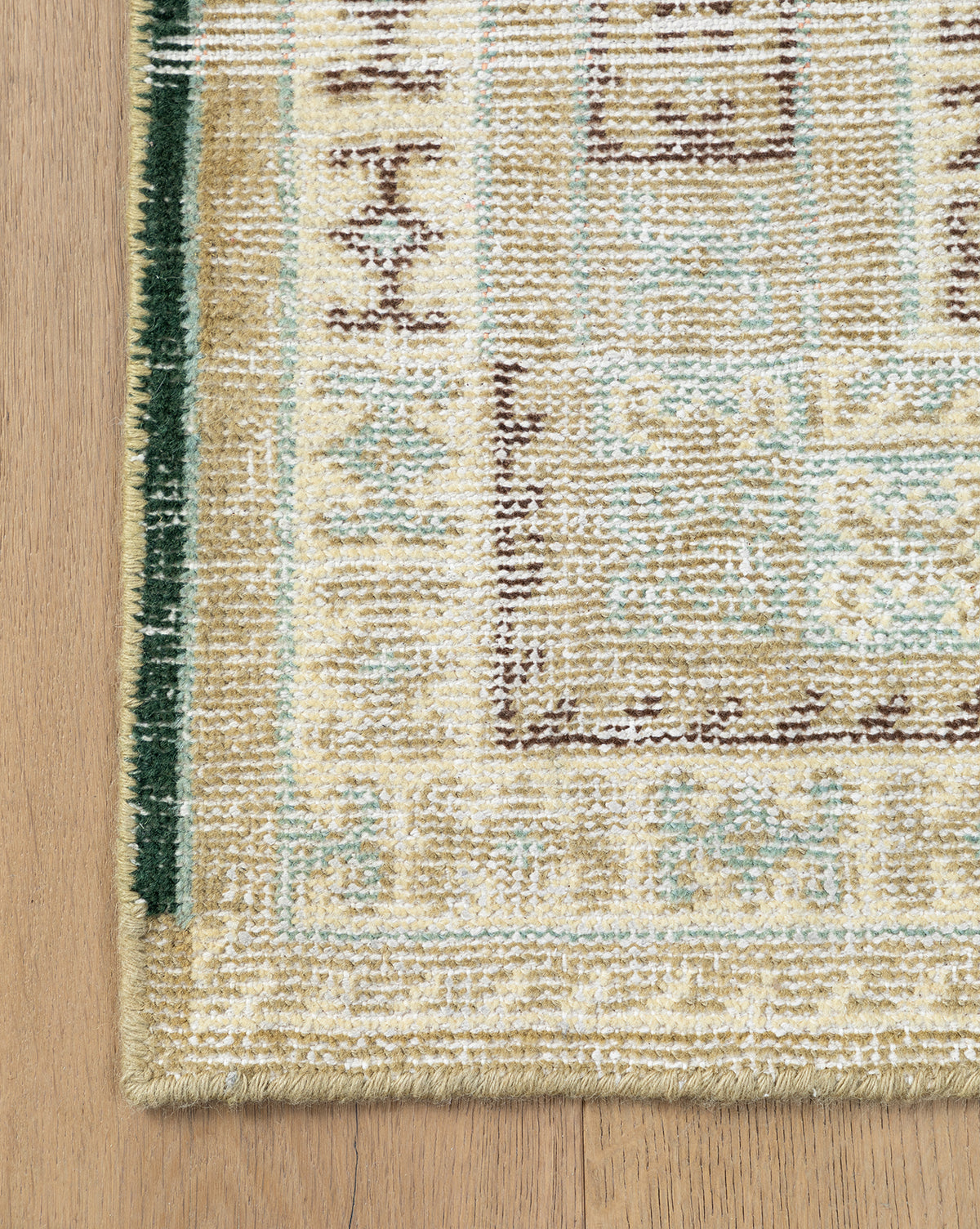 Corner of the Evangeline Hand-Knotted Wool Rug, shown in cream, beige, and pale blue on a wooden floor. This durable wool rug features a green border with intricate geometric patterns, by McGee & Co.