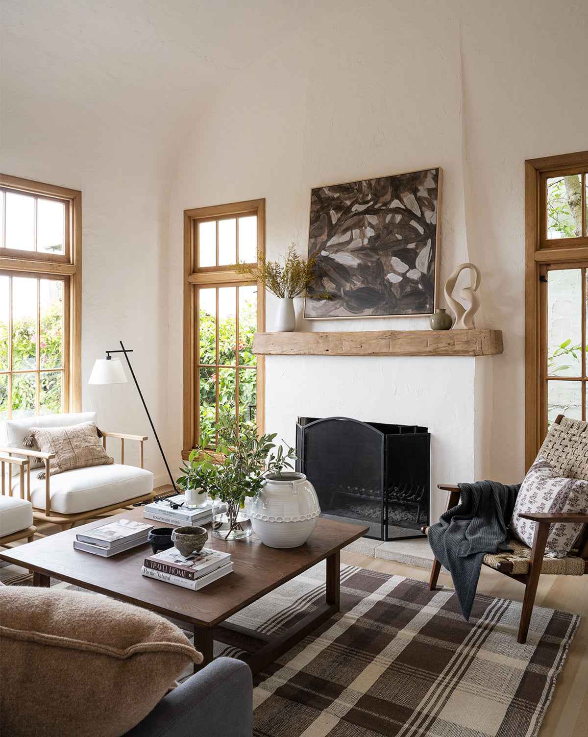 A cozy living room with neutral tones, featuring a wooden coffee table, armchairs, a white sofa, and a fireplace adorned with art. The Orton Handwoven Wool Rug adds texture while large windows fill the space with light and garden views by Vendixs