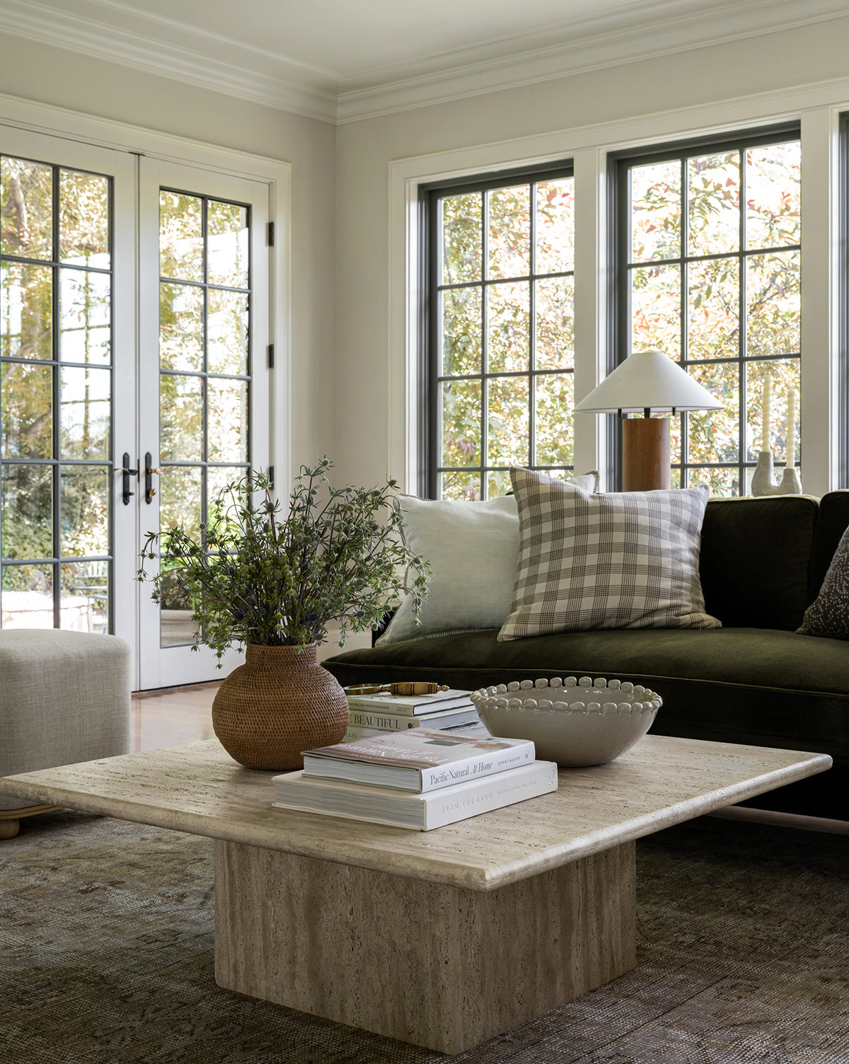 A bright living room with large windows, a dark sofa accented by light and checkered pillows, a square stone coffee table holding books, greenery in a vase, and the Malibu Bowl as décor. Trees are visible outside, by Vendixs