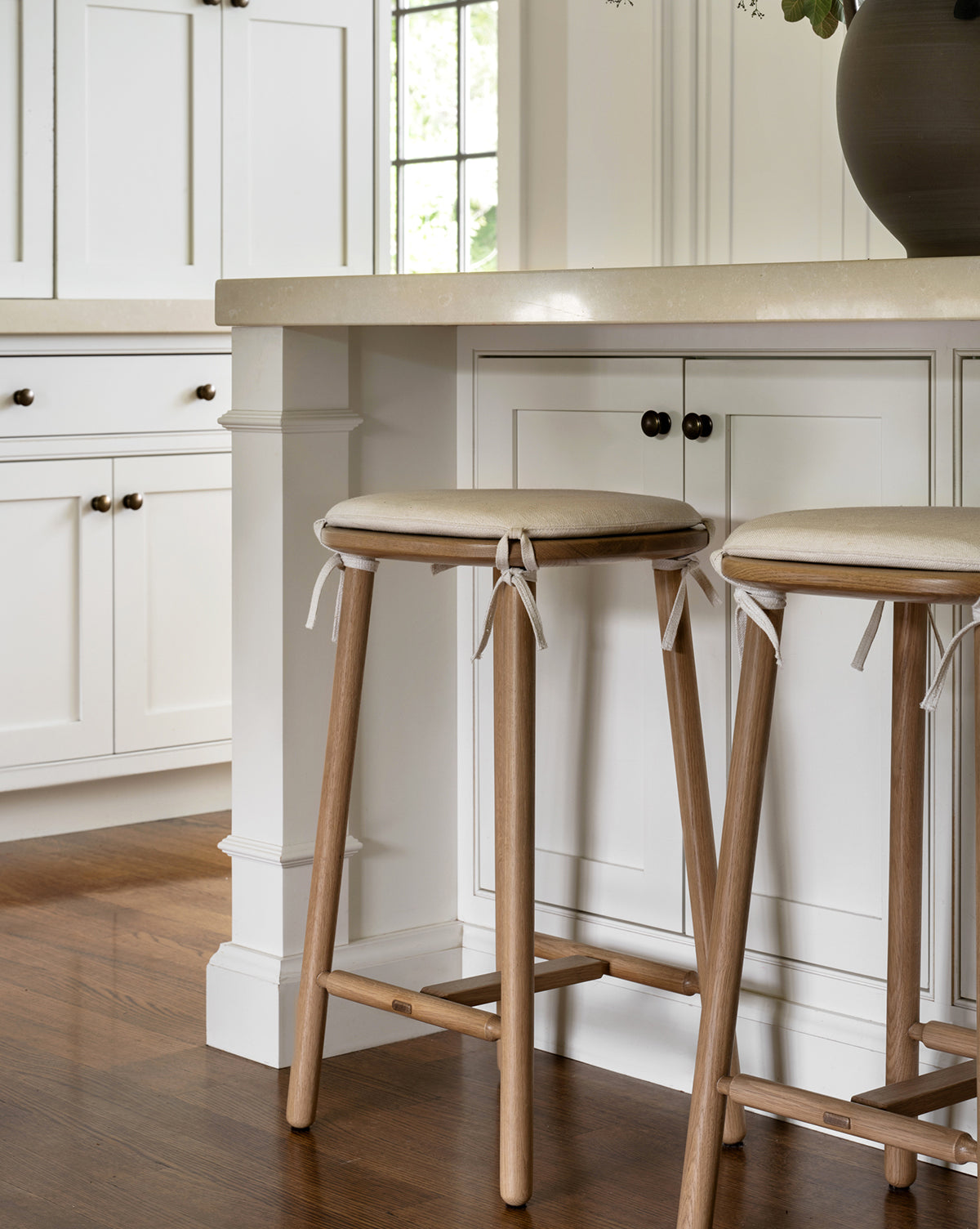 Two Ralph Oak Counter Stools with linen cushions sit at a kitchen counter featuring white cabinetry and a beige countertop, while sunlight through a window highlights the warm wood flooring, by Vendixs