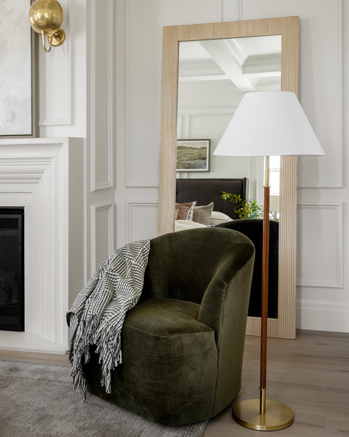 A cozy green velvet chair with a striped throw sits beside the Elijah Floor Lamp, featuring an elegant brass pole, next to a large wooden-framed mirror that reflects a stylish, modern living room with white paneled walls, by Vendixs