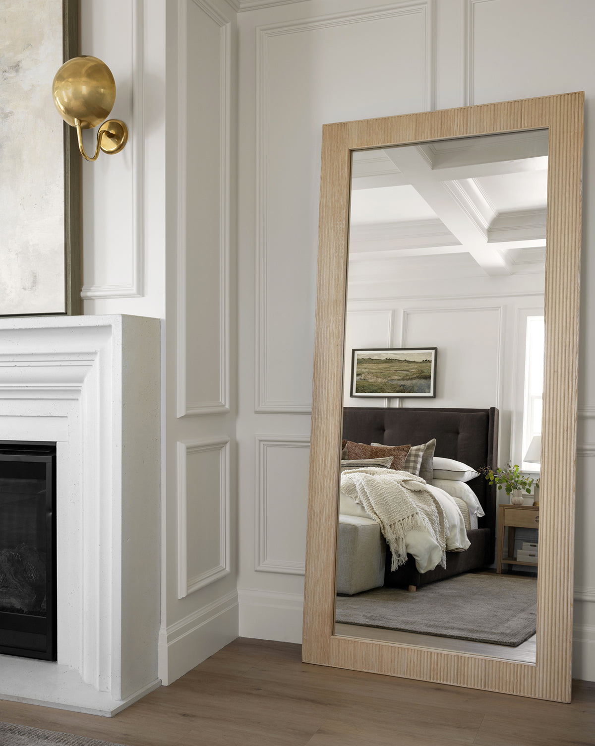 A Rowan Floor Mirror with a ribbed oak frame leans against a white-paneled wall, reflecting a cozy bedroom scene with bed, pillows, nightstand, blanket, and landscape art. A gold wall sconce and part of a fireplace are also in view, by Vendixs