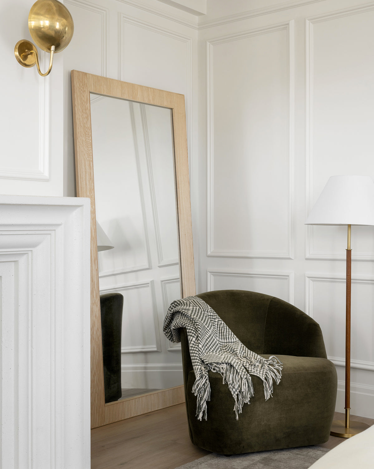 A modern corner with a green velvet Byron Chair draped with a patterned throw, a large wooden-framed mirror, a white floor lamp, white paneled walls, and a brass wall sconce, by Vendixs