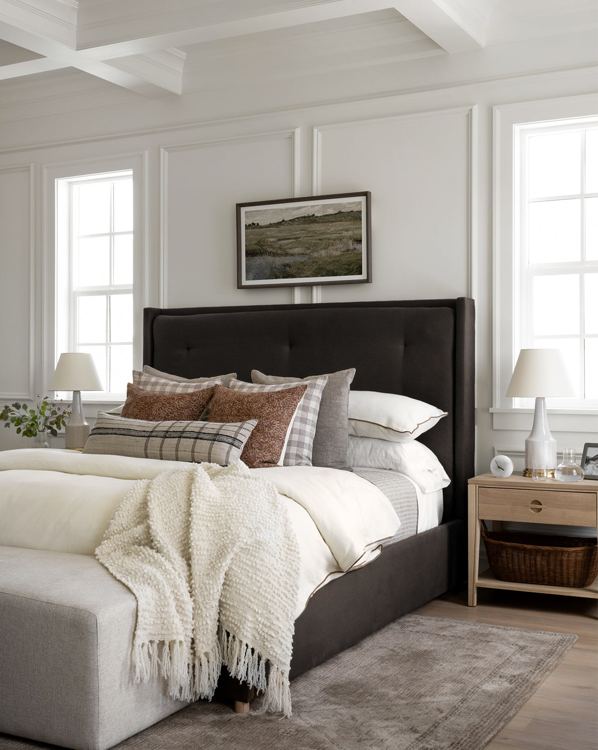 The Ria Bed by Makerspalm, topped with layered pillows and a cream knit blanket, sits in a bright modern bedroom with white paneled walls, large windows, dual bedside tables, and a landscape painting above its tufted headboard by Vendixs