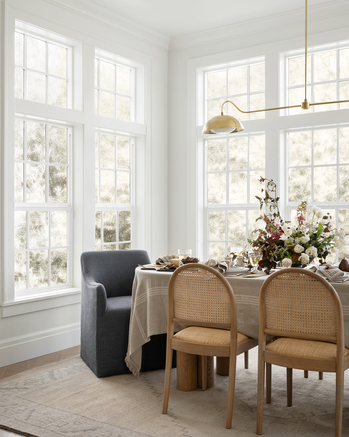 A bright dining area with large windows, a beige tablecloth, four chairs, and a floral centerpiece under a gold light fixture sits atop the Pembroke Hand-Knotted Wool Rug for vintage-inspired charm, by Vendixs