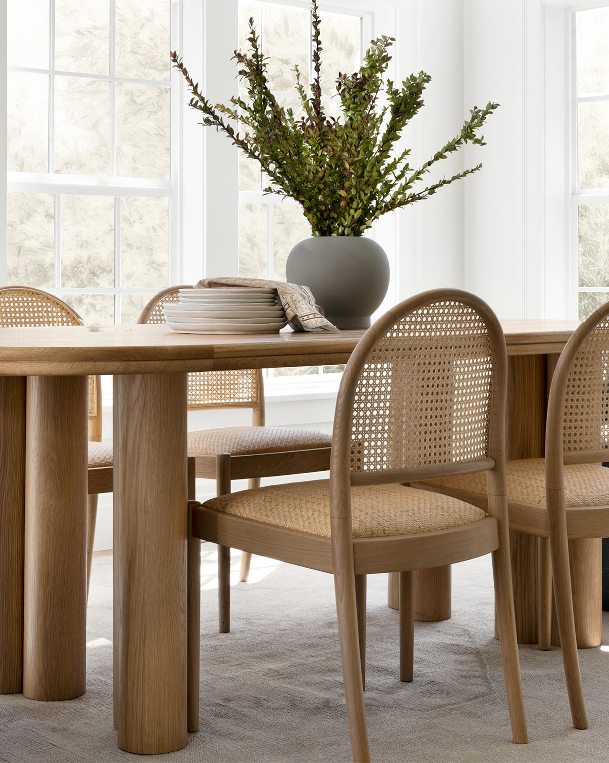 A light wood dining table with four Hadden Natural Dining Chairs featuring cane backs sits in a bright room. A gray vase with green branches and a stack of plates with napkins decorate the table near large windows, by Vendixs