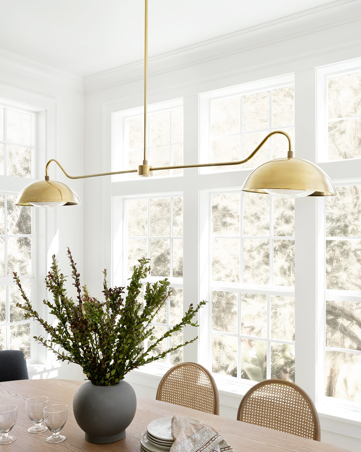 A modern dining area with a light wood table, rattan chairs, a black vase with greenery, and the Aldorno Linear Chandelier by Makerspalm, set before large sunlit windows that frame a snowy outdoor view. Vendixs