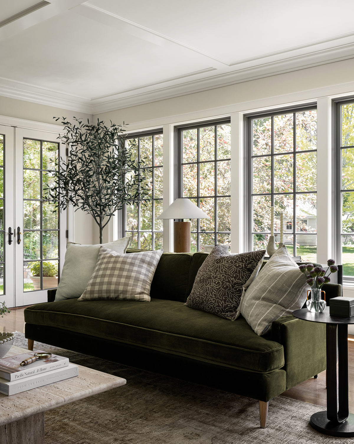 A modern living room with large windows, a green Makerspalm Evan Sofa 84 with patterned pillows, a side table and lamp, potted tree, and coffee table with books—all bathed in natural light. Vendixs
