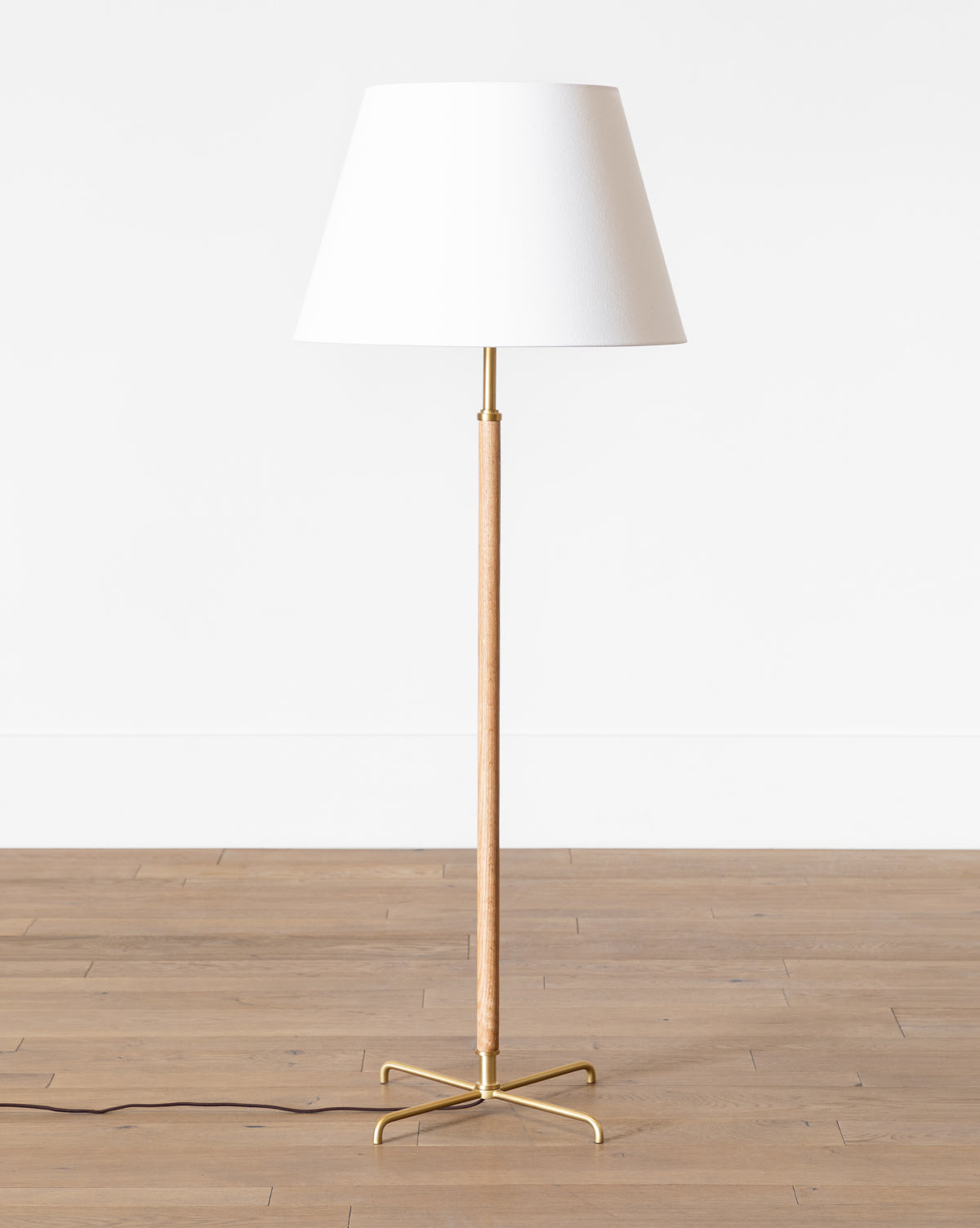 The Makerspalm Florence Floor Lamp features a slim wooden stand, gold metal base, and elegant linen shade, standing on a wooden floor against a plain white wall. Vendixs