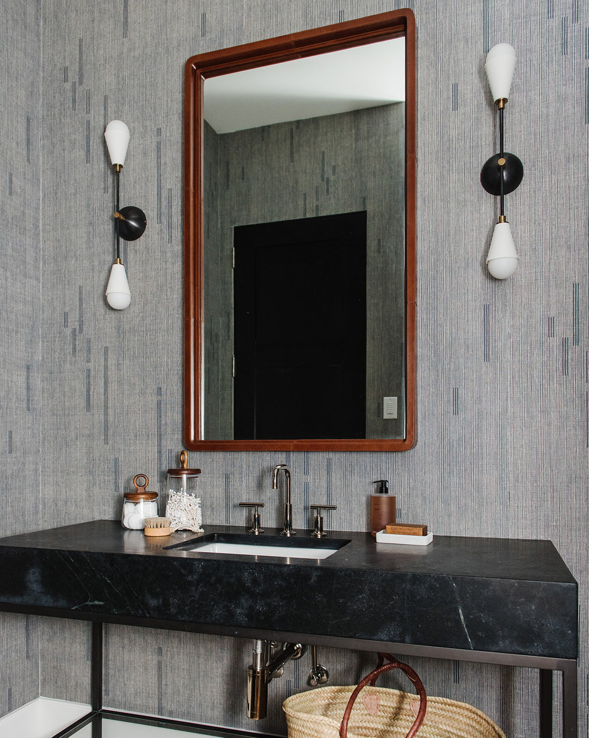 A modern bathroom features a black countertop, rectangular sink, and chrome faucet. Above hangs the Franklin Leather Mirror, flanked by black and white sconces. A woven basket sits under the sink, by Vendixs