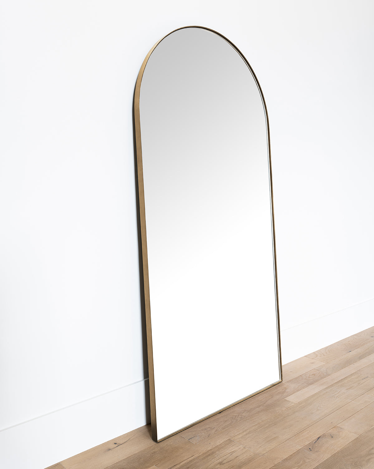 The Gala Floor Mirror features a large reflective surface and a slim gold frame, leaning against a white wall on light wood flooring. Its modern, minimalist design enhances any room with elegance and simplicity, by Vendixs