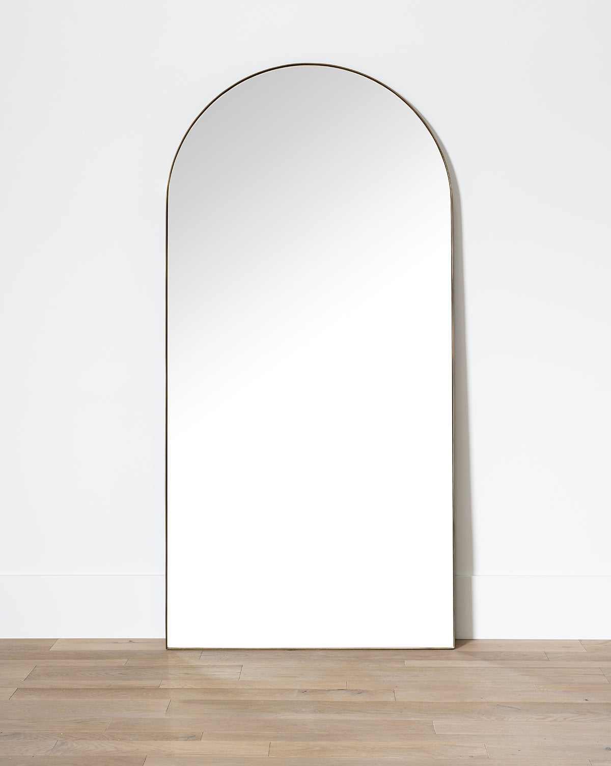 The Gala Floor Mirror, featuring a slim frame, is shown leaning against a white wall on a light wood floor, its reflection capturing the simplicity of the wall across from it, by Vendixs