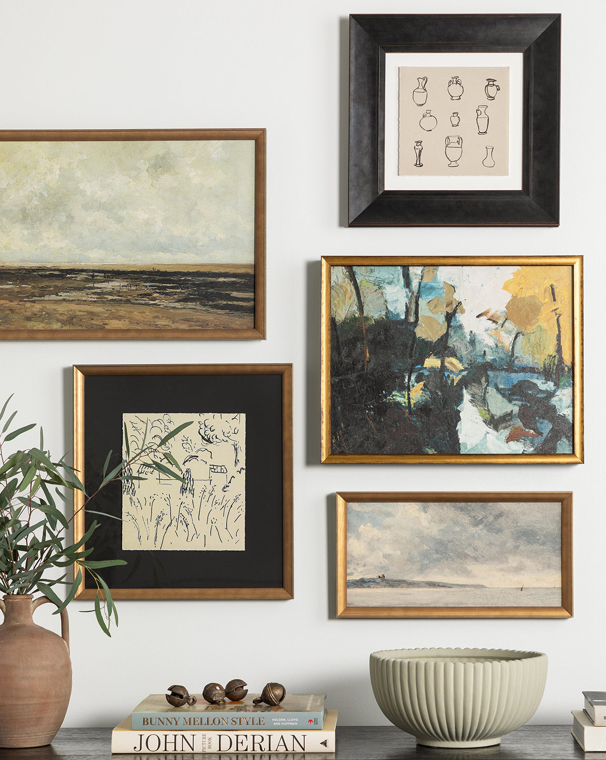 A gallery wall featuring five framed artworks, such as the Gray Seas abstract landscape, hangs above a table styled with a potted plant, decorative bowl, and stacked books for a balanced, moody look.