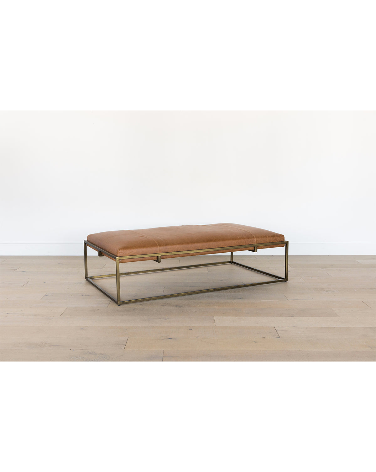 The Harlow Leather Coffee Table, featuring a rectangular brown aniline-dyed leather cushion and a modern brass-finished frame, sits on a light wood floor against a plain white wall, by Vendixs