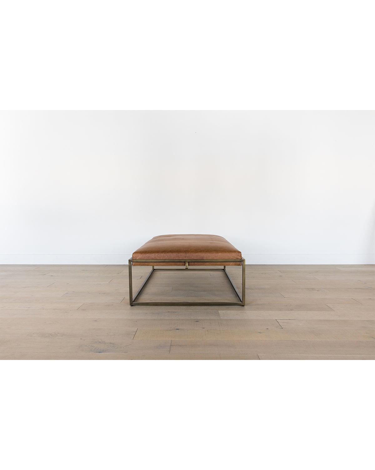 The Harlow Leather Coffee Table, featuring a sleek brass-finished frame and aniline-dyed leather top, sits on a light wooden floor against a plain white wall, by Vendixs