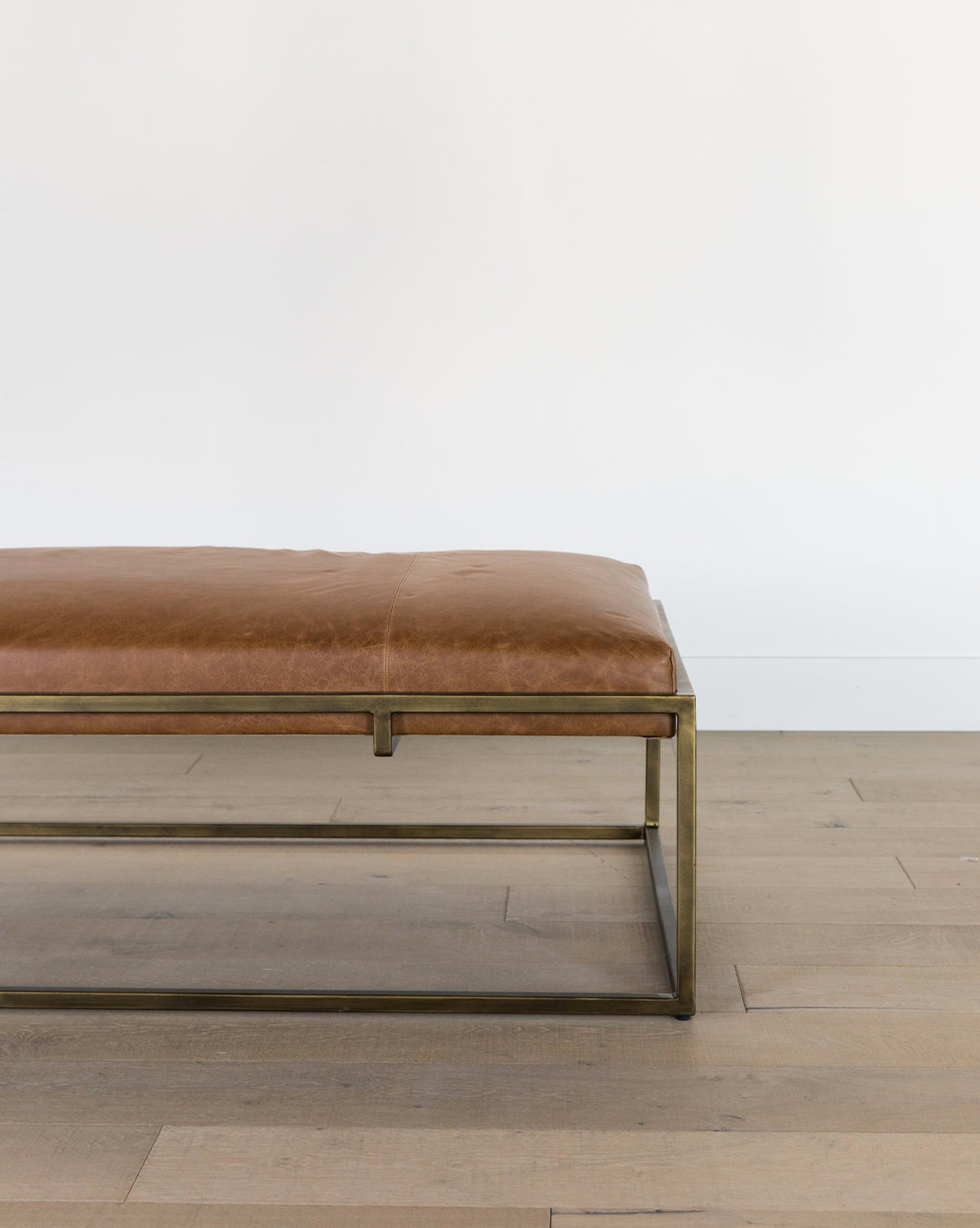The Harlow Leather Coffee Table, featuring an aniline-dyed leather cushion and a sleek brass-finished frame, sits on a light wooden floor against a white wall. Only the right half of the table is visible, by Vendixs