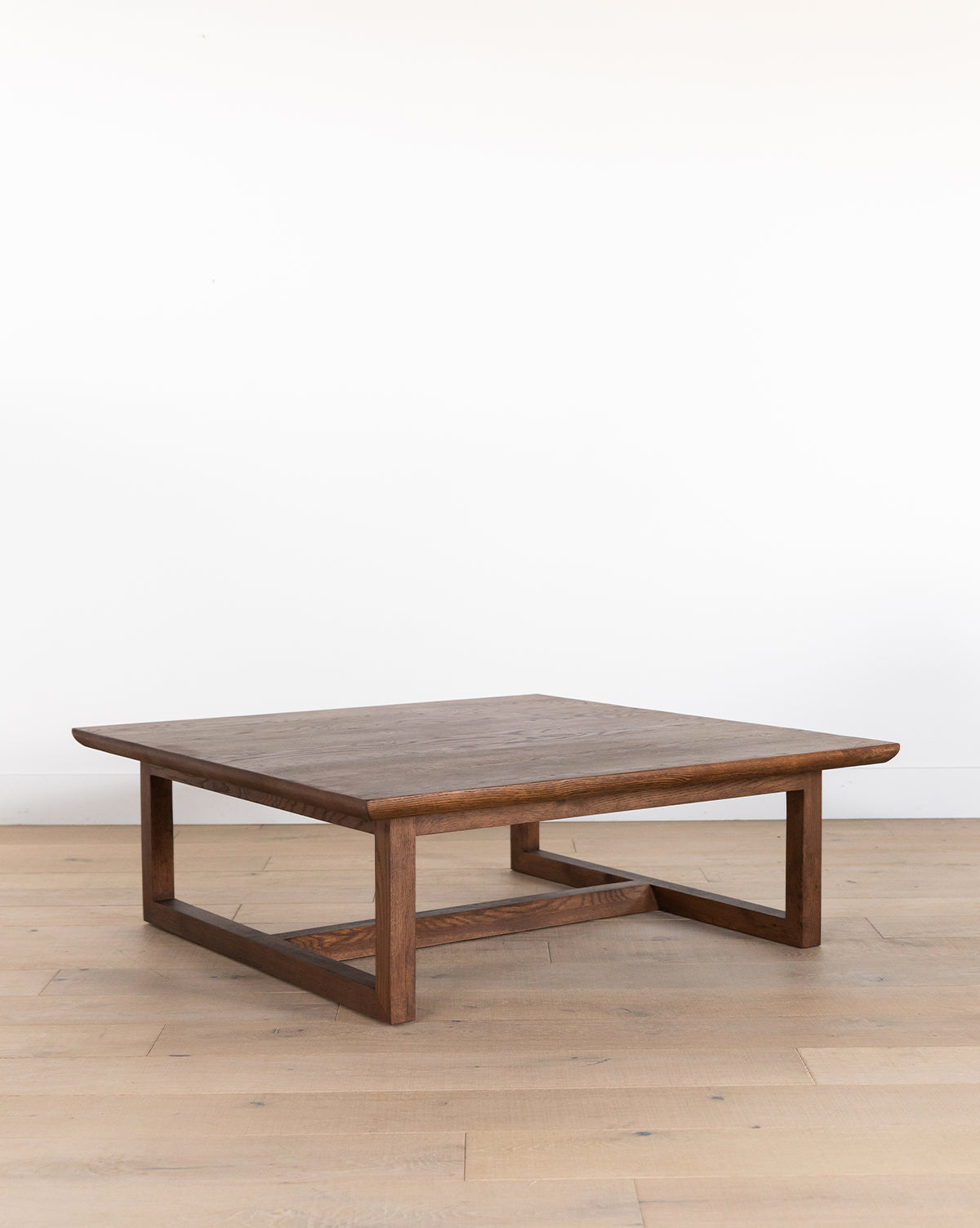 The Herrin Coffee Table is a modern, square coffee table made from solid oak, with a flat top and open rectangular legs, shown on a light wood floor in front of a plain white wall, by Vendixs