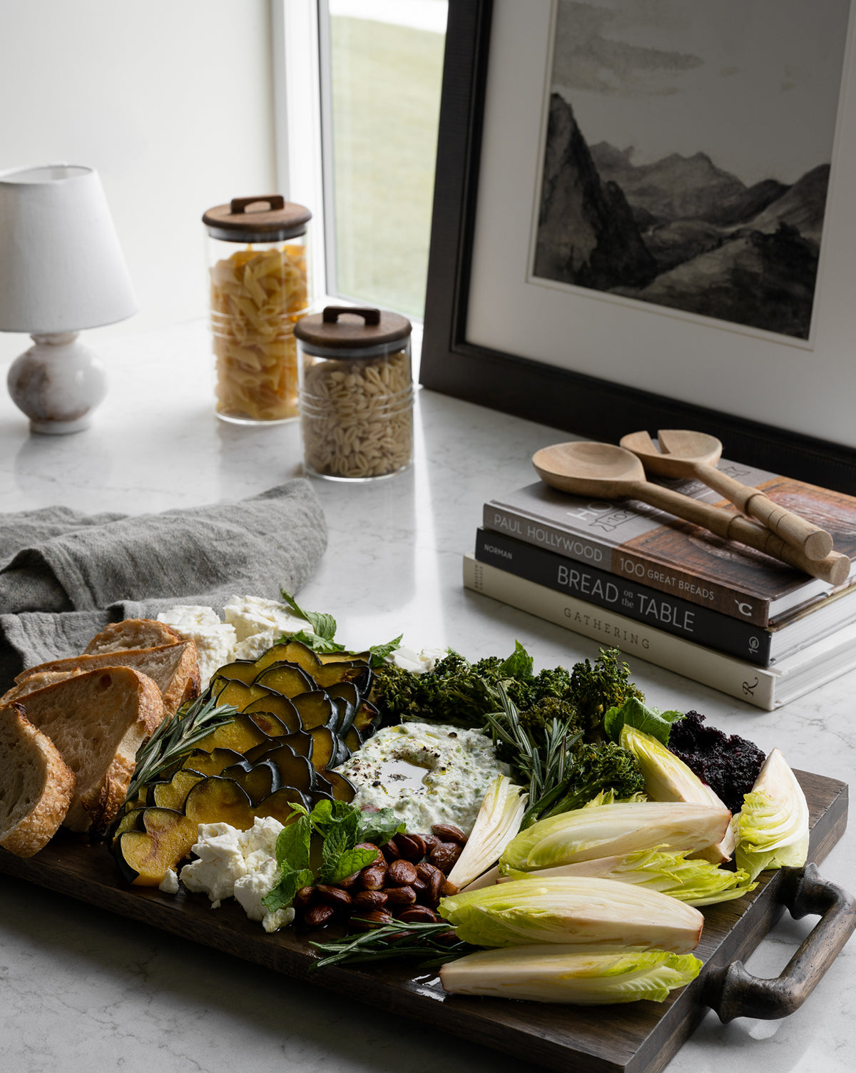 A charcuterie board with grilled squash, broccoli, nuts, cheese, and endive is served on the Fordham Board near toasted bread, books, wooden utensils, pasta jars, a lamp, and framed art by the window by Vendixs