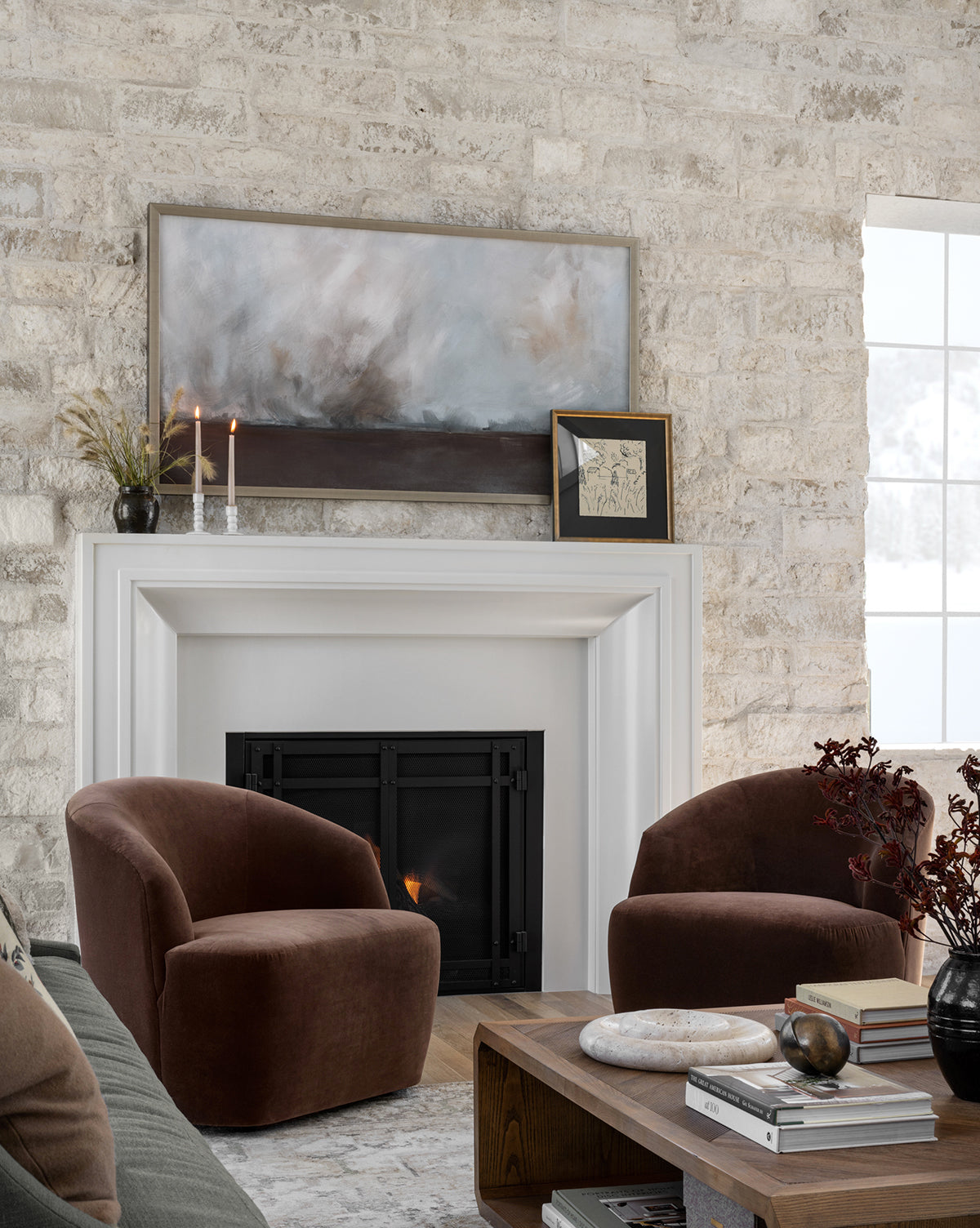 Cozy living room with two Byron Chairs in custom fabric by a white fireplace, abstract art and candles on the mantel, a wooden coffee table with books and decor, and a large snowy window view, by McGee & Co.