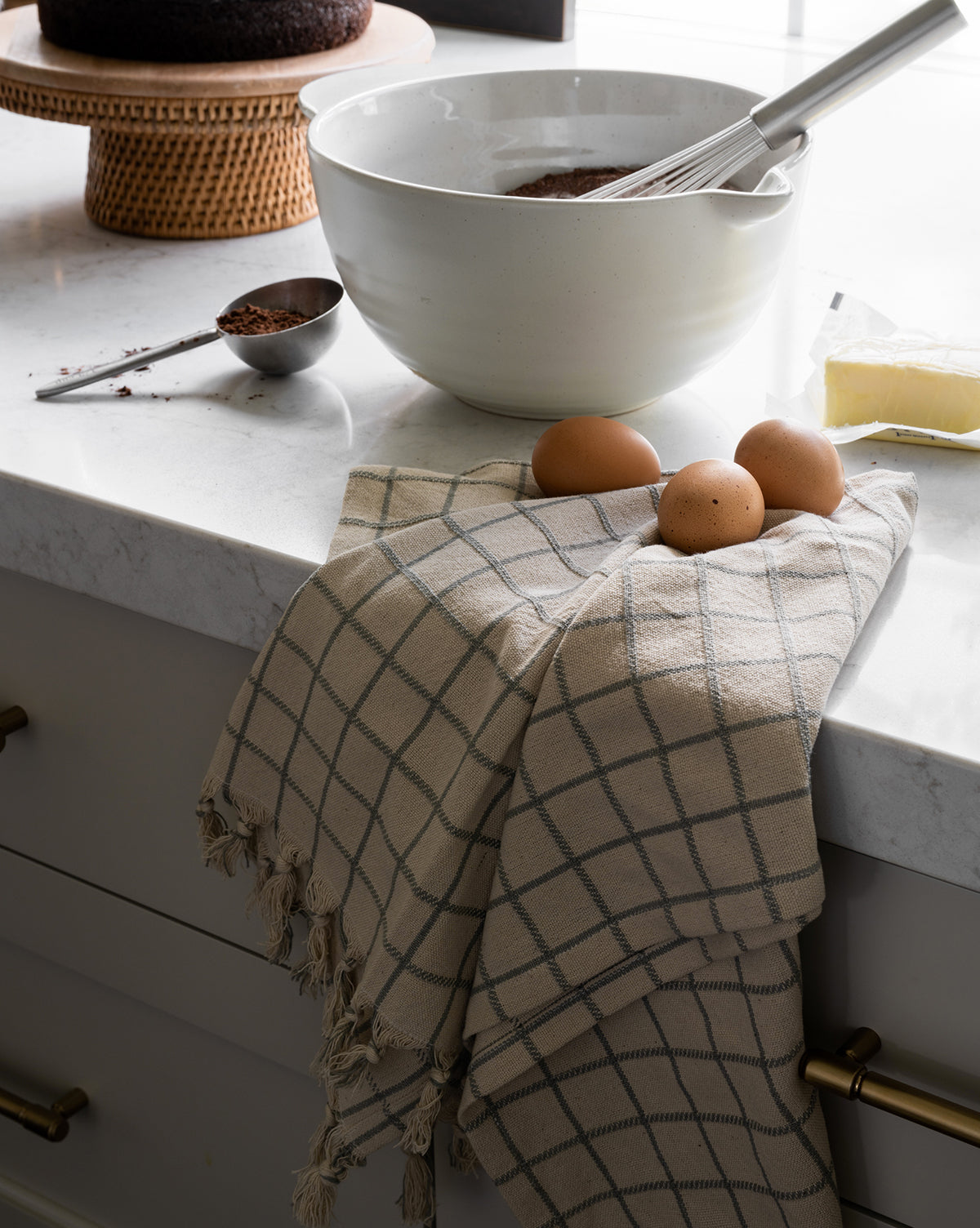A bowl with a whisk, baking ingredients, three brown eggs on the Peabody Cotton Tea Towel (Set of 2), a stick of butter, and a chocolate cake on a stand sit on a white marble counter—perfect kitchen inspiration, by Vendixs