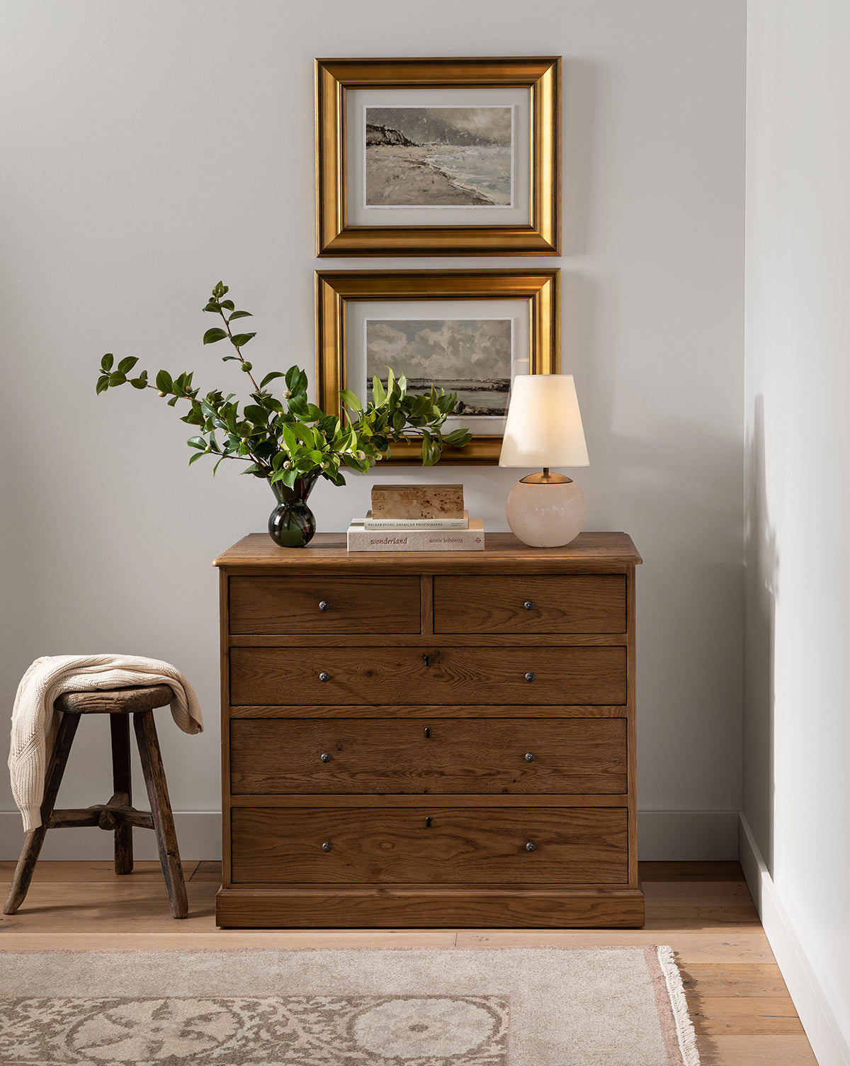 A wooden dresser with six drawers sits against a white wall, topped with a green leafy vase, stacked books, and the Terri Round Table Lamp with its alabaster stone base. Two framed landscape paintings hang above. A small wooden stool is on the left, by McGee & Co.