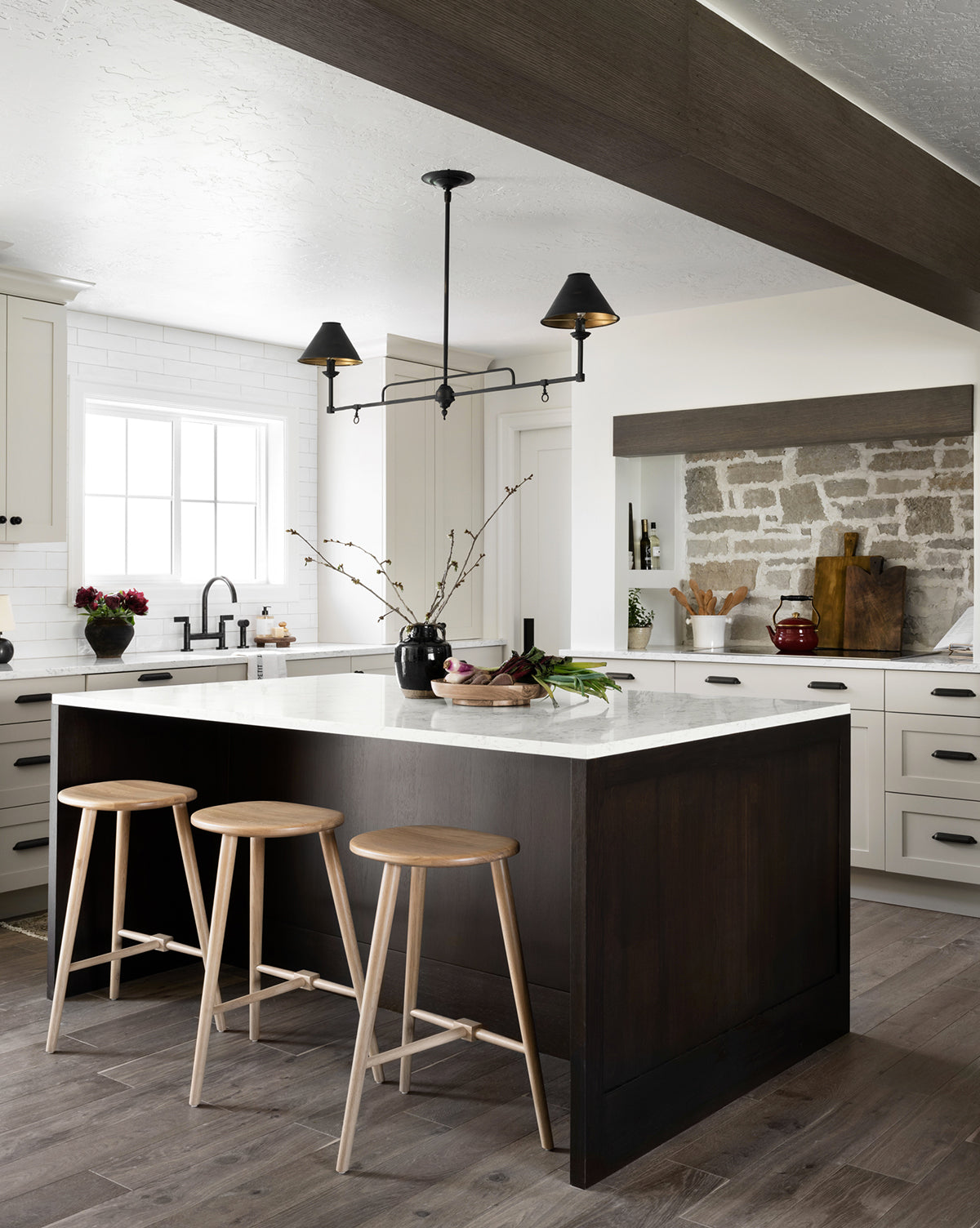 Modern kitchen featuring three Fielder Counter Stools in light oak at a large dark wood island with white countertops, neutral cabinets, a farmhouse sink, exposed brick backsplash, and minimalist pendant lights above by Vendixs