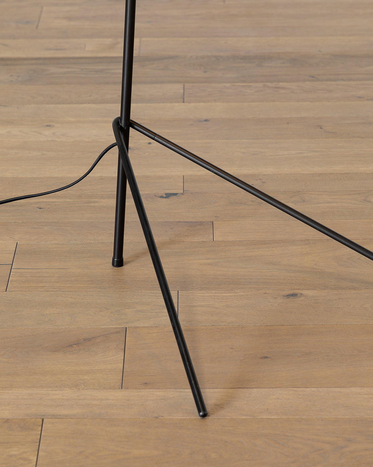 The Johanson Woven Floor Lamp features black metal tripod legs standing on light wood flooring, with a visible black power cord extending from the lamp, by Vendixs