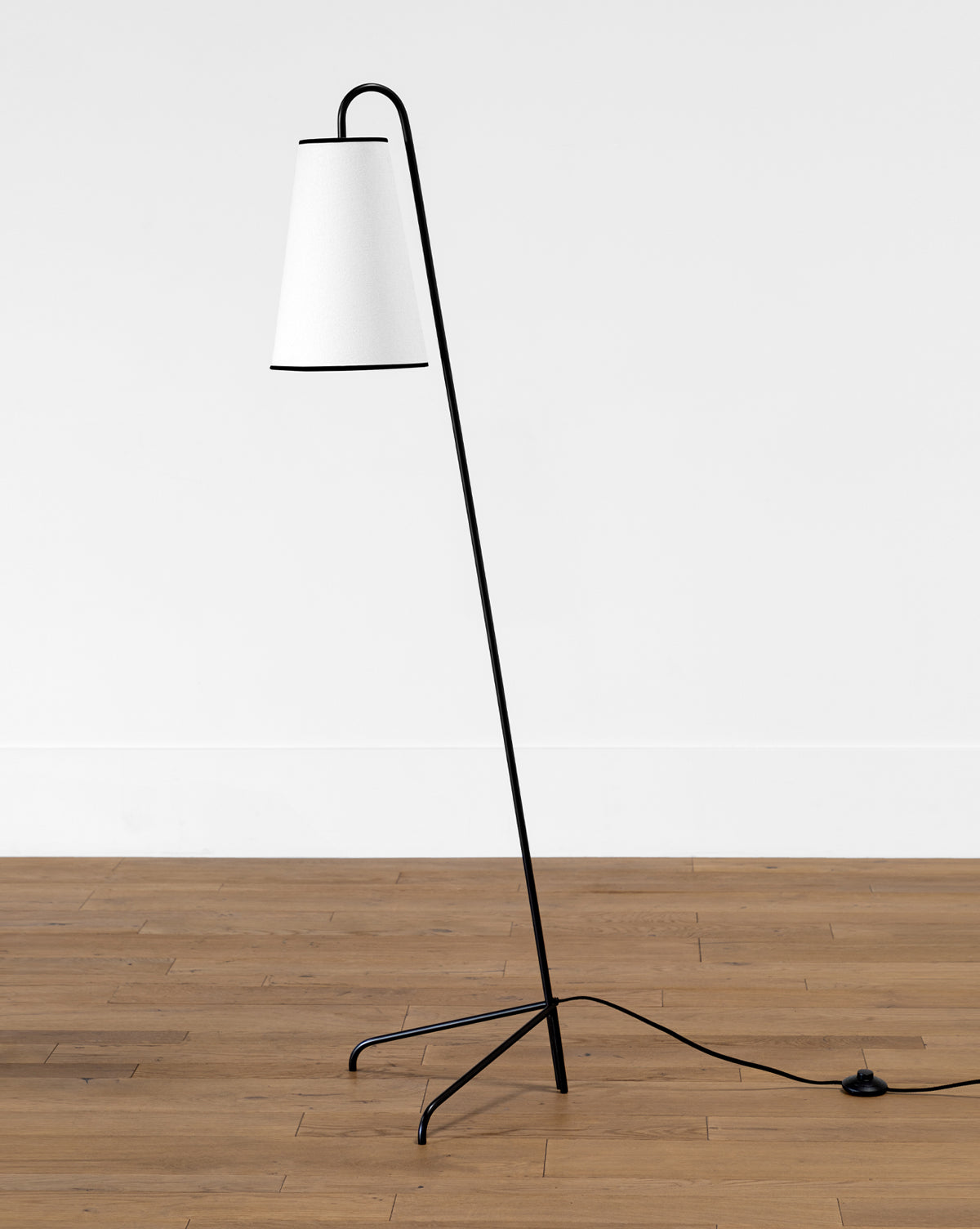 The Makerspalm Jonas Floor Lamp has a slender black metal frame, angled stem, white cone-shaped linen shade, and three-pronged legs, with a visible black power cord for a modern look. Vendixs