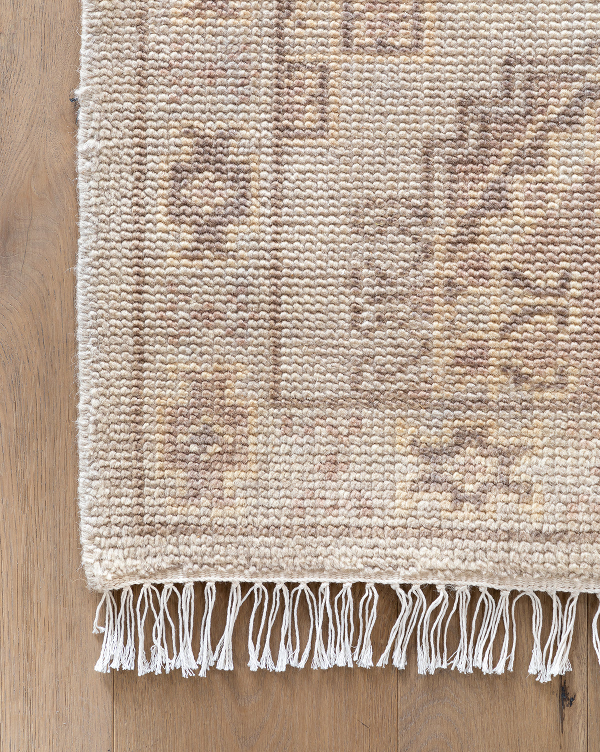 A close-up of the corner of the beige Kenna Hand-Knotted Wool Rug, featuring a subtle geometric pattern and fringed edges, shown on a light wood floor, by Vendixs