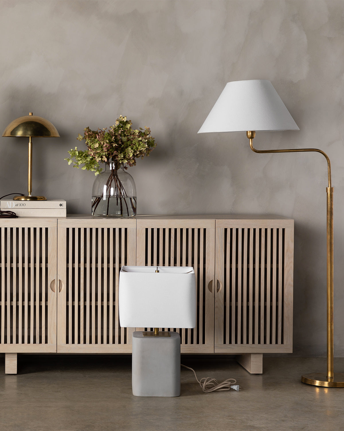 Minimalist interior featuring a light wooden sideboard, brass table lamp, hydrangeas in a glass vase, books, and the Wainwright Swoop Floor Lamp placed on the floor against a gray wall, by Vendixs