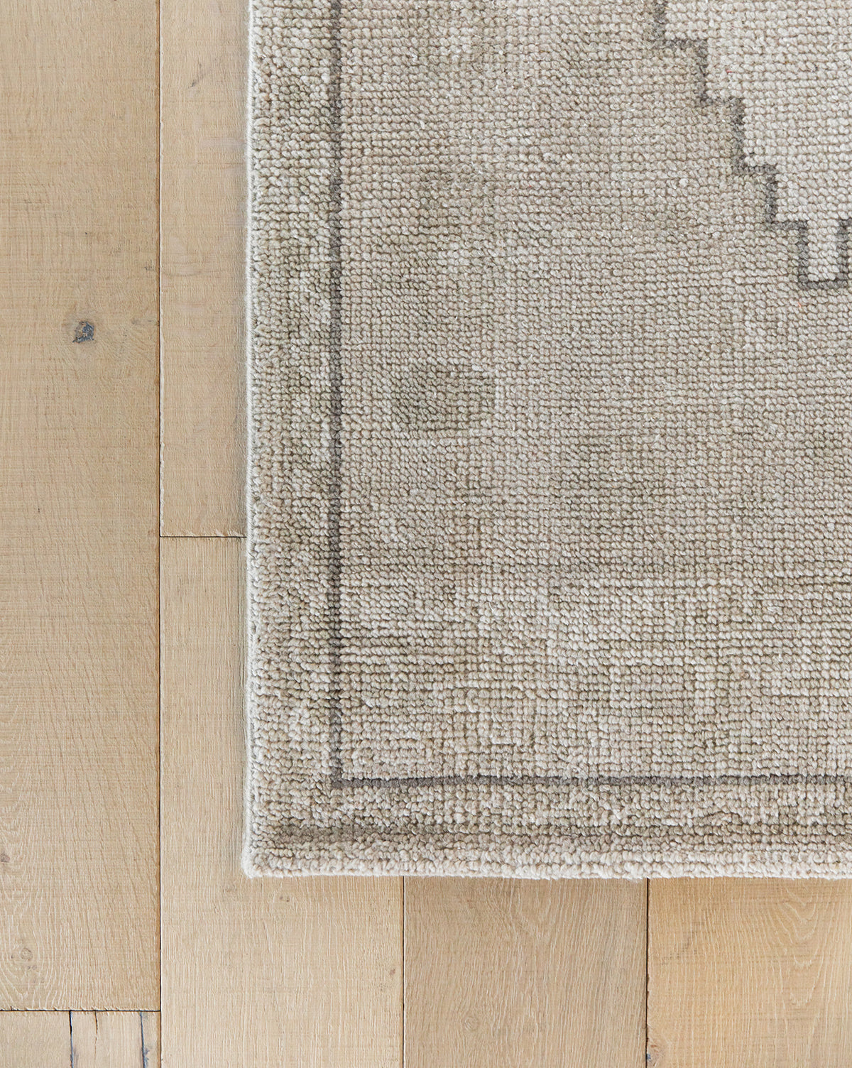 A close-up of the Pembroke Hand-Knotted Wool Rug, featuring a light tan textured transitional design with a subtle geometric border, shown on light wood flooring with its corner and part of the wooden floor visible, by Vendixs